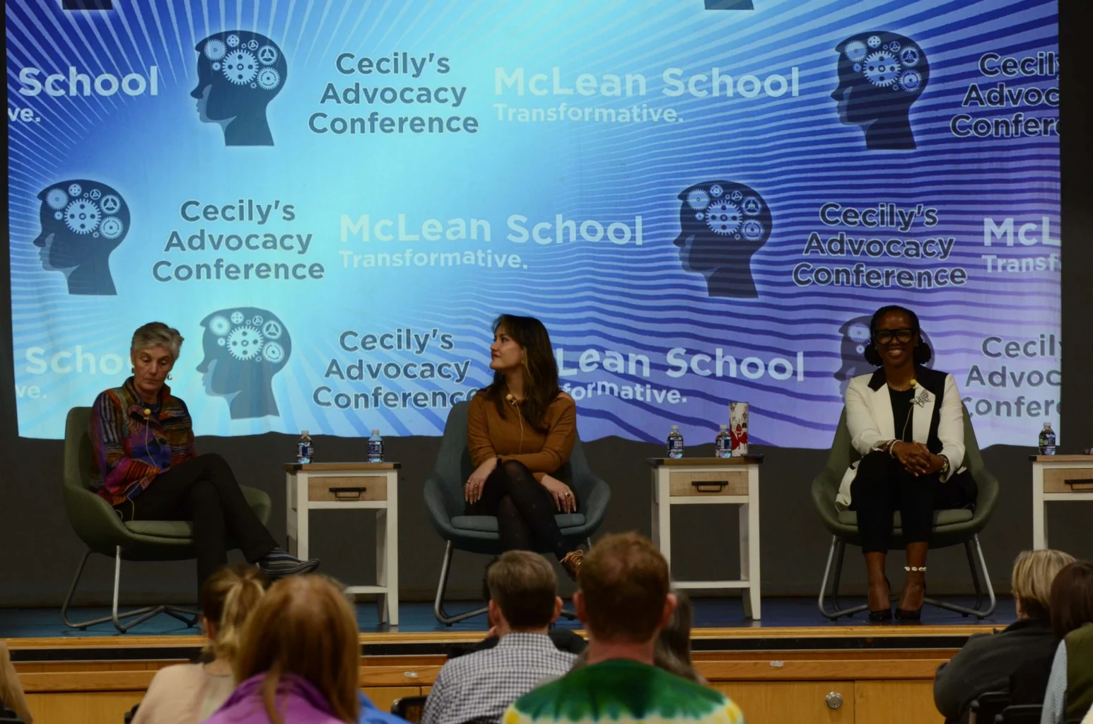 Three women sit on stage in chairs during a panel discussion at Cecily's Advocacy Conference, with a McLean School banner displayed behind them. Audience members are visible in the foreground.