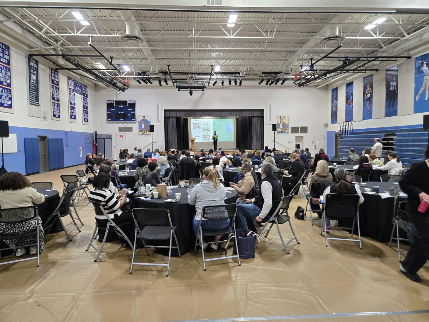 People seated at round tables in a gymnasium attend a presentation, with a speaker on stage and slides projected on a large screen.