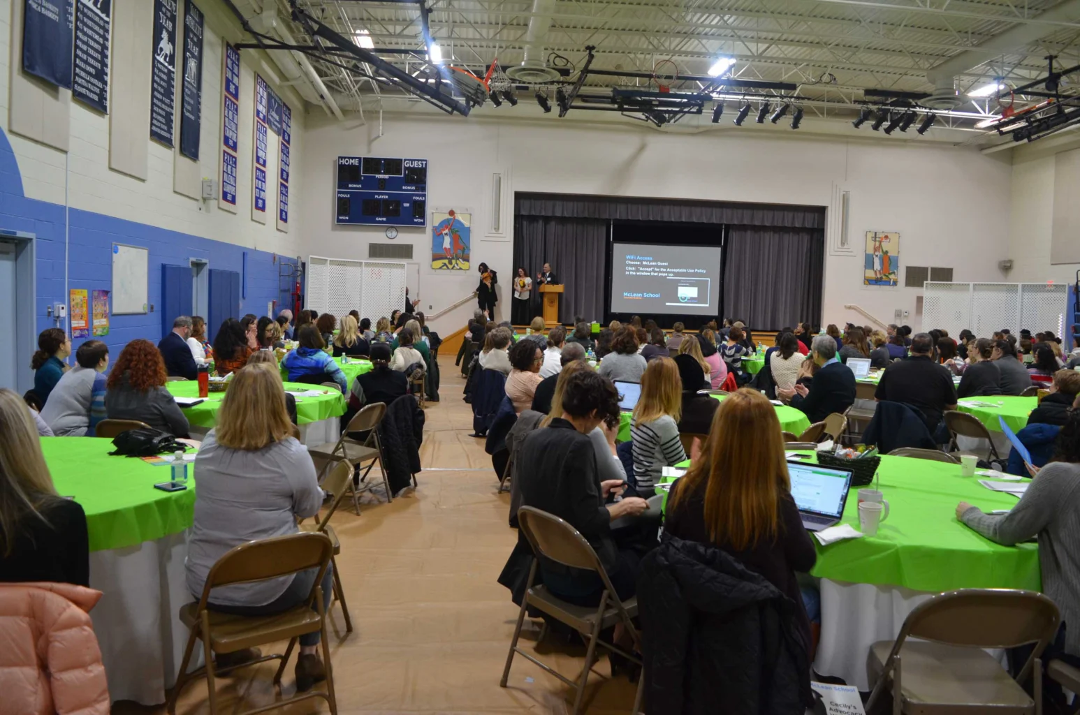 People seated at round tables in a gymnasium watch two presenters speak on stage with a projected slide behind them.