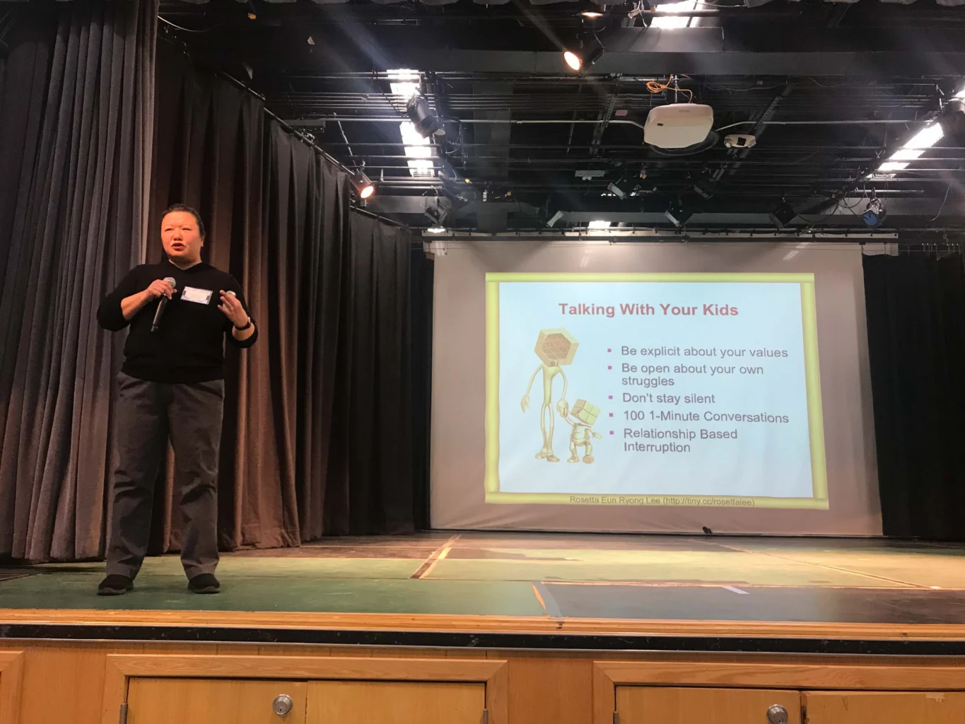 A person stands on a stage speaking, next to a large screen displaying a slide titled "Talking With Your Kids" with bullet points on communication tips.