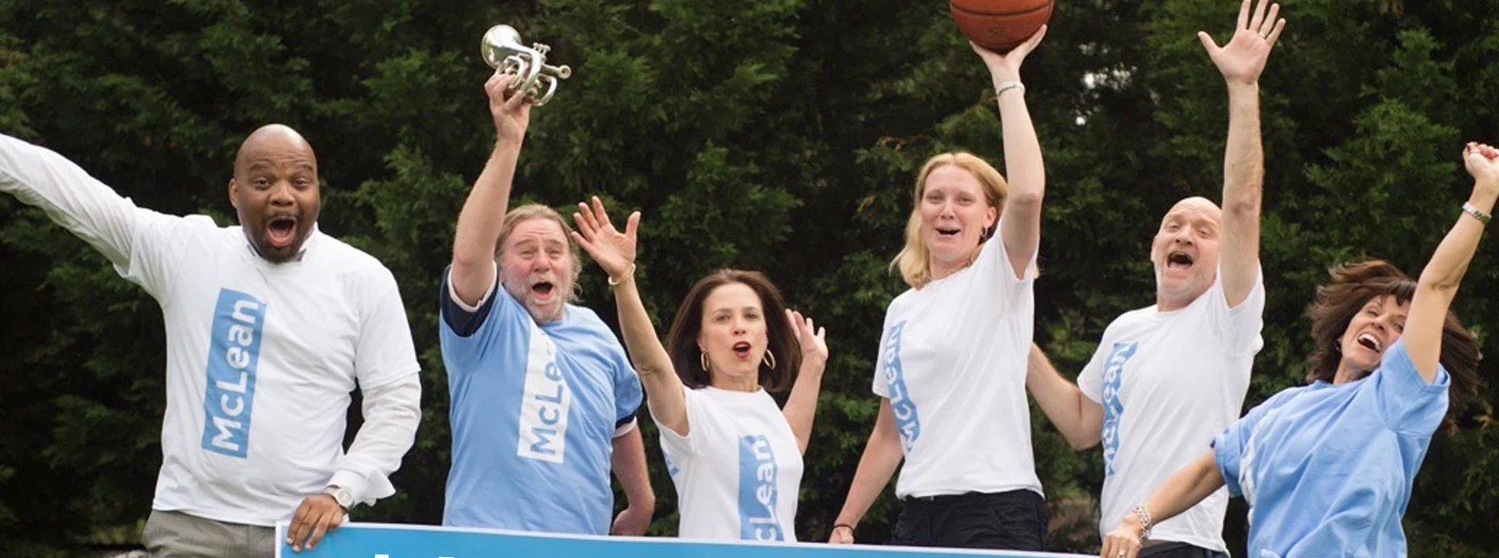 Six adults in McLean t-shirts stand outdoors, holding a trophy and basketball, raising their arms and smiling, with trees in the background.