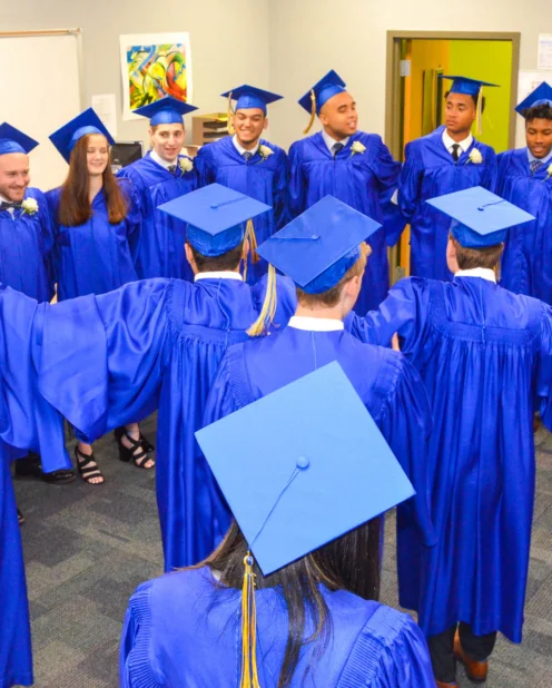 A group of students in blue graduation gowns and caps stand in a circle in a classroom, some holding hands, preparing for a ceremony.