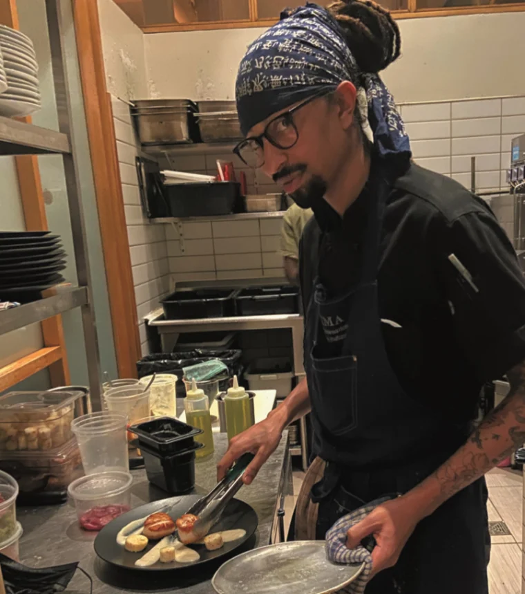 A chef wearing a blue bandana and glasses prepares food on a hot plate in a restaurant kitchen.