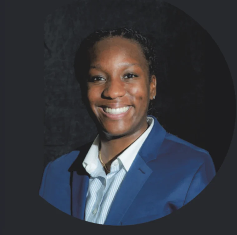 A person wearing a blue blazer and striped shirt smiles in front of a dark background.