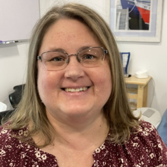 A woman with shoulder-length blonde hair and glasses smiles at the camera, wearing a maroon patterned blouse, in an indoor setting with office items in the background.
