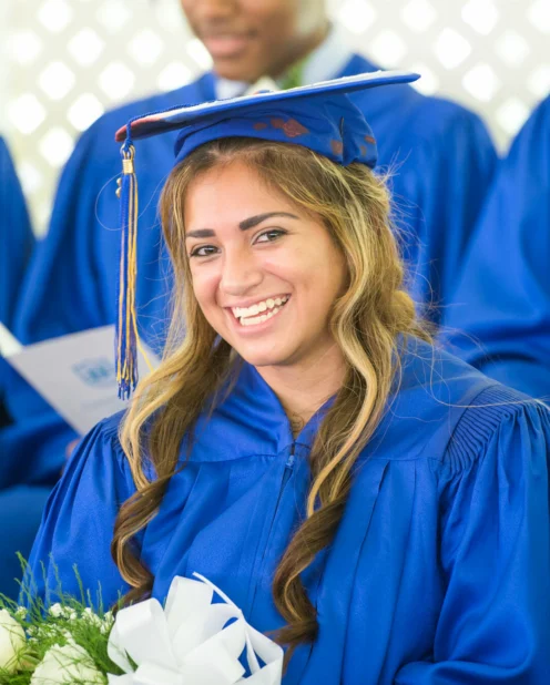 A young woman in a blue graduation gown and cap smiles while holding a bouquet. Other graduates in blue gowns are visible in the background.