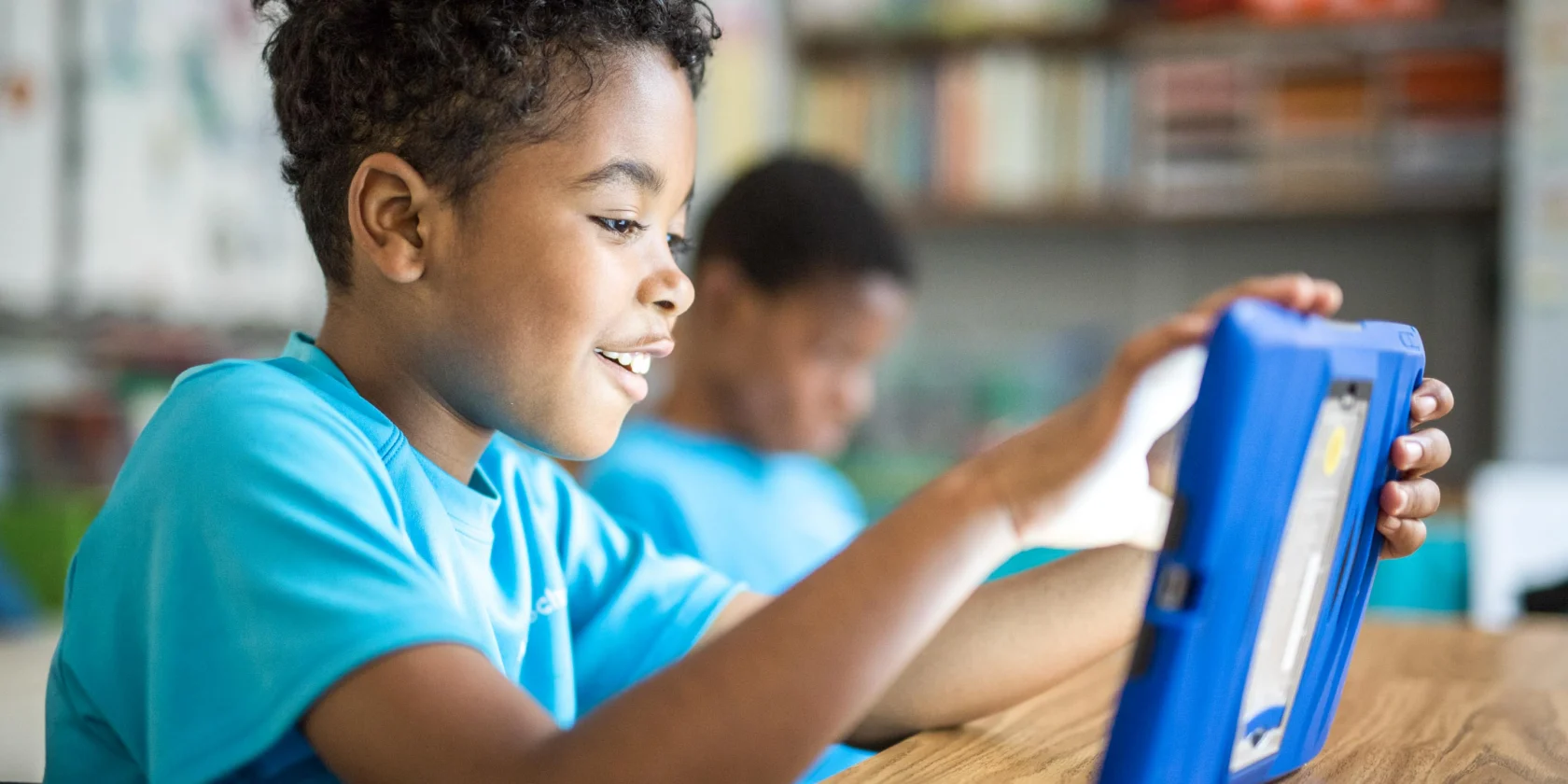 A boy in a blue shirt sits at a desk, smiling and using a tablet in a classroom. Another student in the background also uses a tablet.