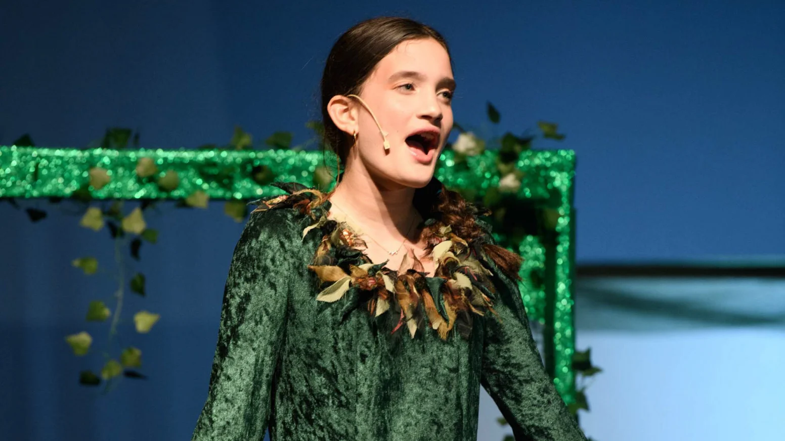 A young girl wearing a green costume with a leaf collar sings on stage, speaking or performing with a headset microphone.