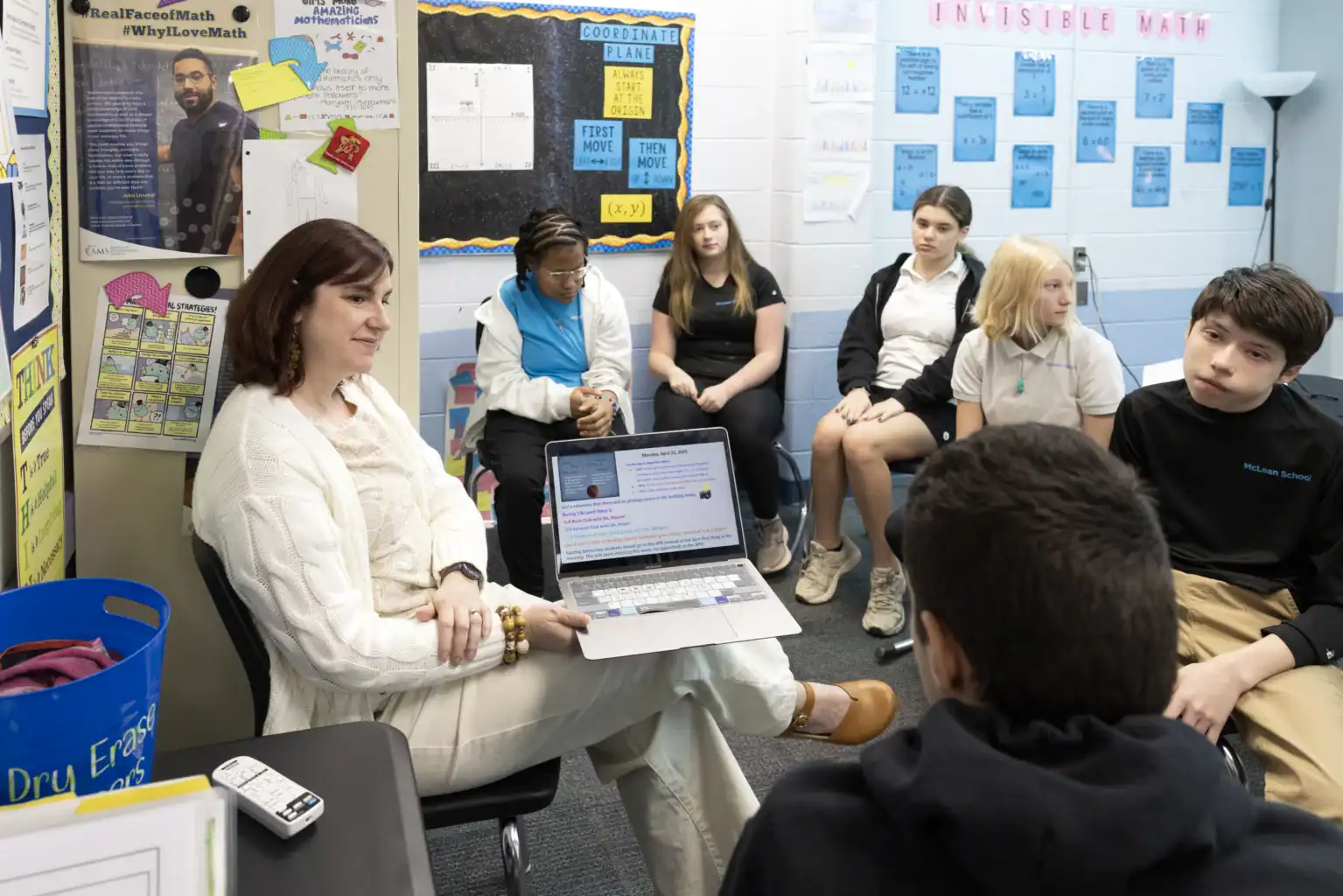 A teacher sits with a laptop on her lap, leading a discussion with a group of middle school students seated in a classroom decorated with educational posters.