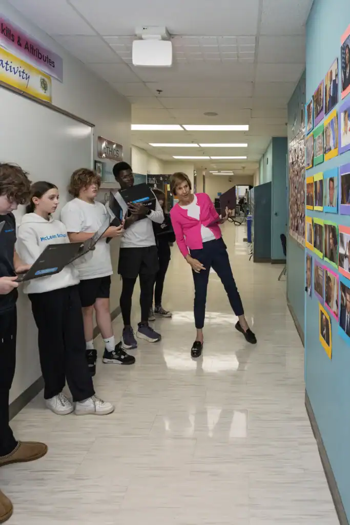A woman in a pink blazer talks to a group of students holding laptops in a hallway lined with colorful photos and posters.