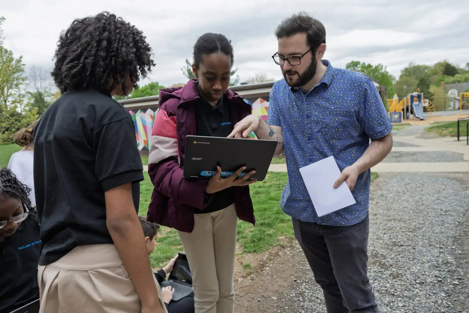 A man holding papers assists two students with a laptop outdoors near a playground and colorful mural.