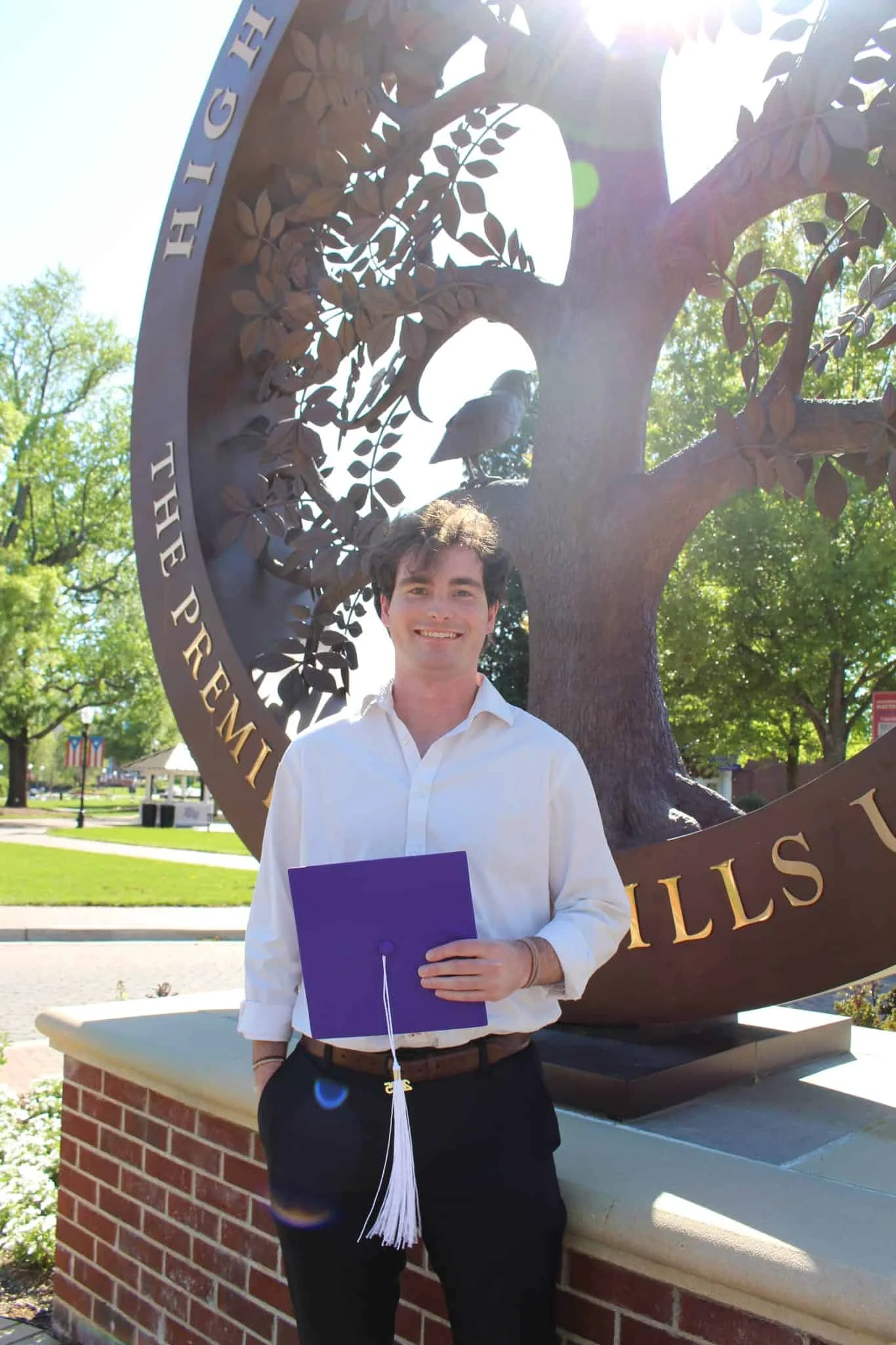A young man in a white shirt holds a purple diploma holder and stands in front of a large tree sculpture with gold lettering on a sunny day.