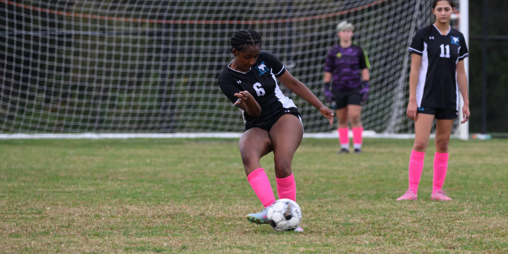 A soccer player in a black uniform and pink socks kicks a ball on a field, with two teammates and a goal in the background.