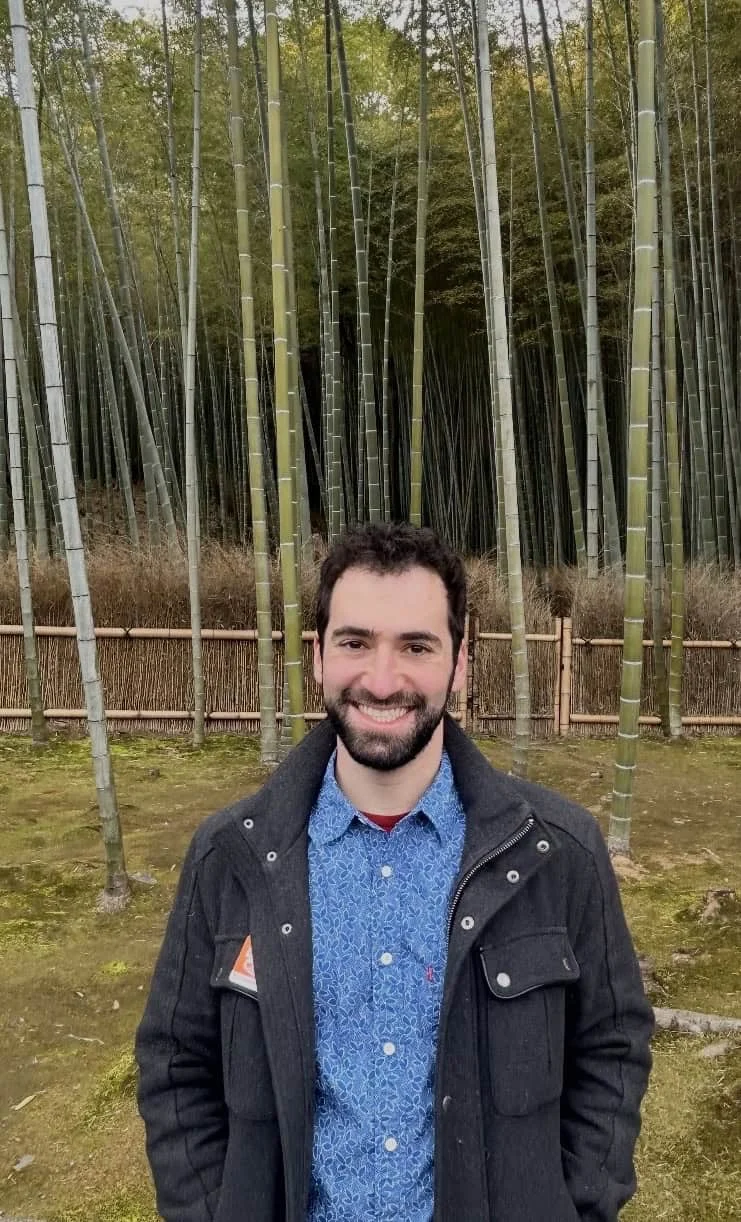 A man with a beard stands smiling in front of tall bamboo stalks and a wooden fence, wearing a blue patterned shirt and a dark jacket.