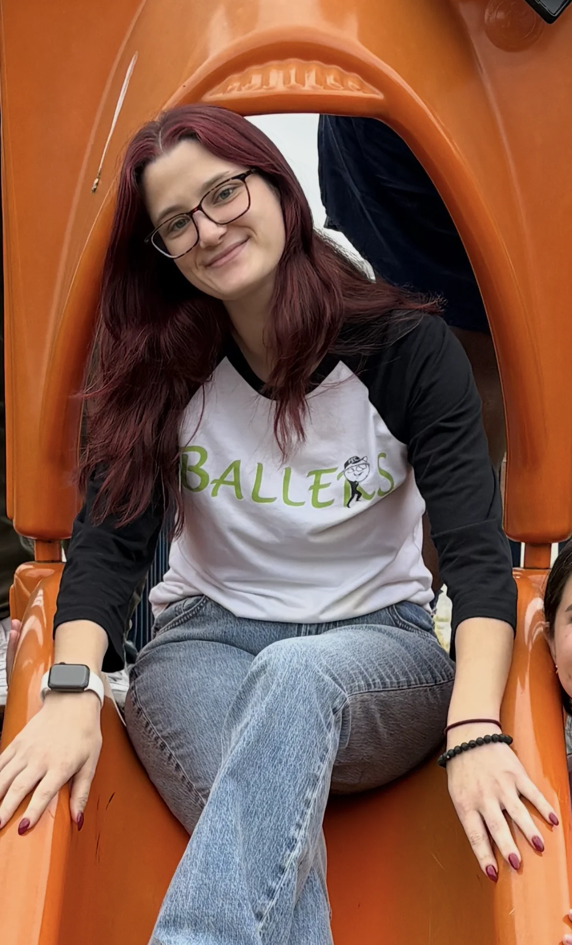 Bella Gleim, Kindergarten. Person with long red hair and glasses sits on an orange slide, wearing a white and black “Ballers” shirt, blue jeans, and sneakers, smiling at the camera.