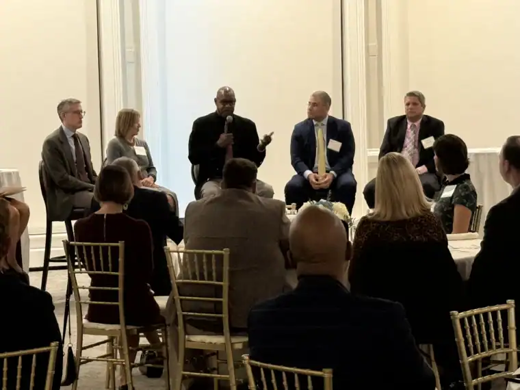 Five panelists speak to an audience in a formal setting, seated in a row on stage, with attendees listening and taking notes on gold chairs.